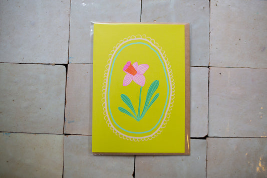 Greeting card with a pink daffodil design on a yellow background, placed on a tiled floor.