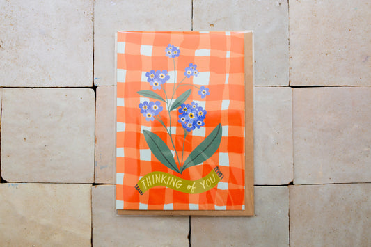 Card floral design and 'Thinking of You' text on an orange checkered background, placed on a stone surface.