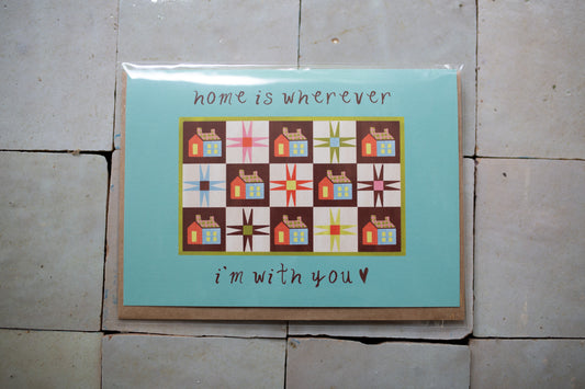 Greeting card with quilt design and text 'home is wherever I'm with you' on a tiled floor.