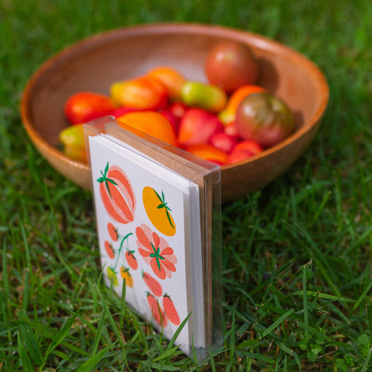 A box of illustrated tomato-themed cards stands upright on green grass next to a wooden bowl filled with assorted tomatoes.