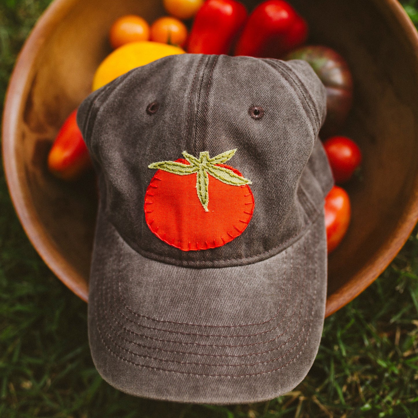A dark brown baseball cap with a red tomato patch and green leaf embroidery on the front, resting on a wooden bowl filled with colorful tomatoes and peppers on a grassy background.