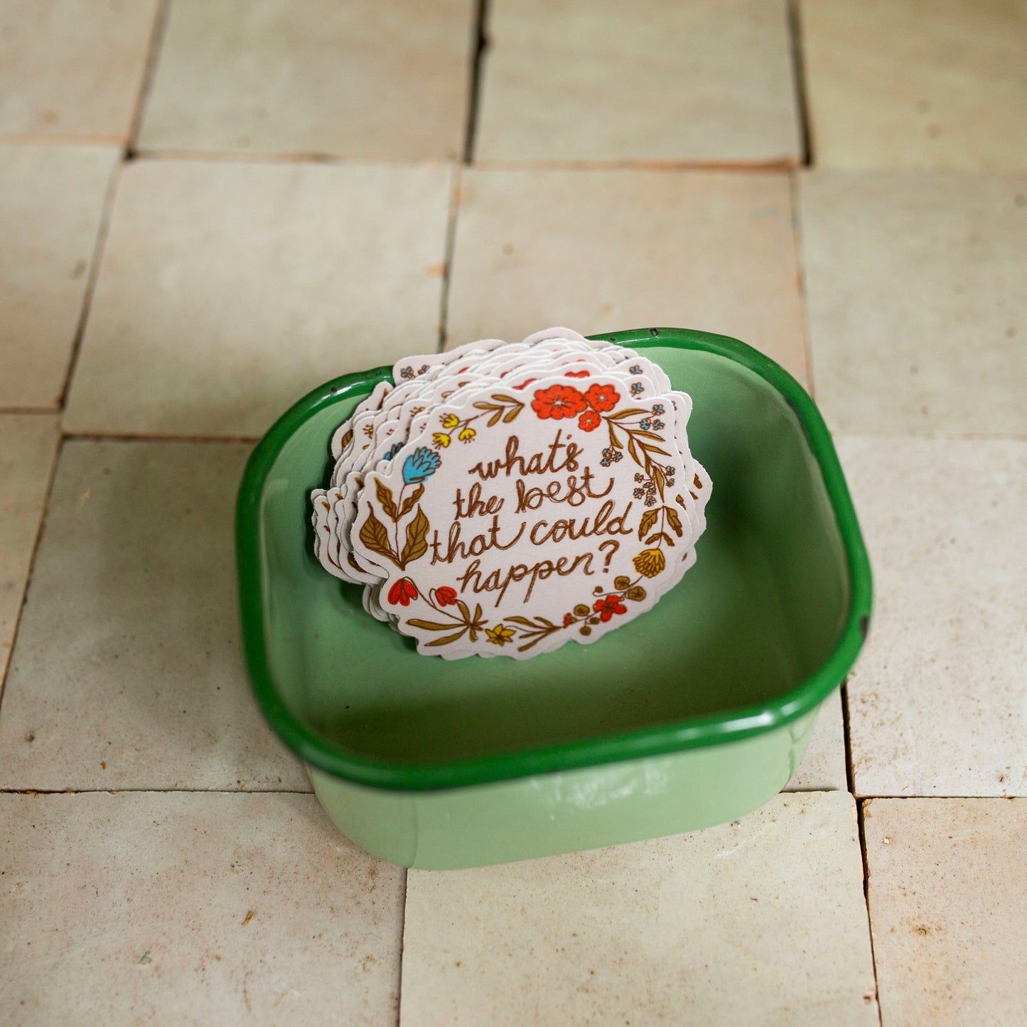 A stack of white paper stickers with floral borders and the phrase 'what's the best that could happen?' printed in brown script, placed inside a small green square dish on a beige tiled floor.