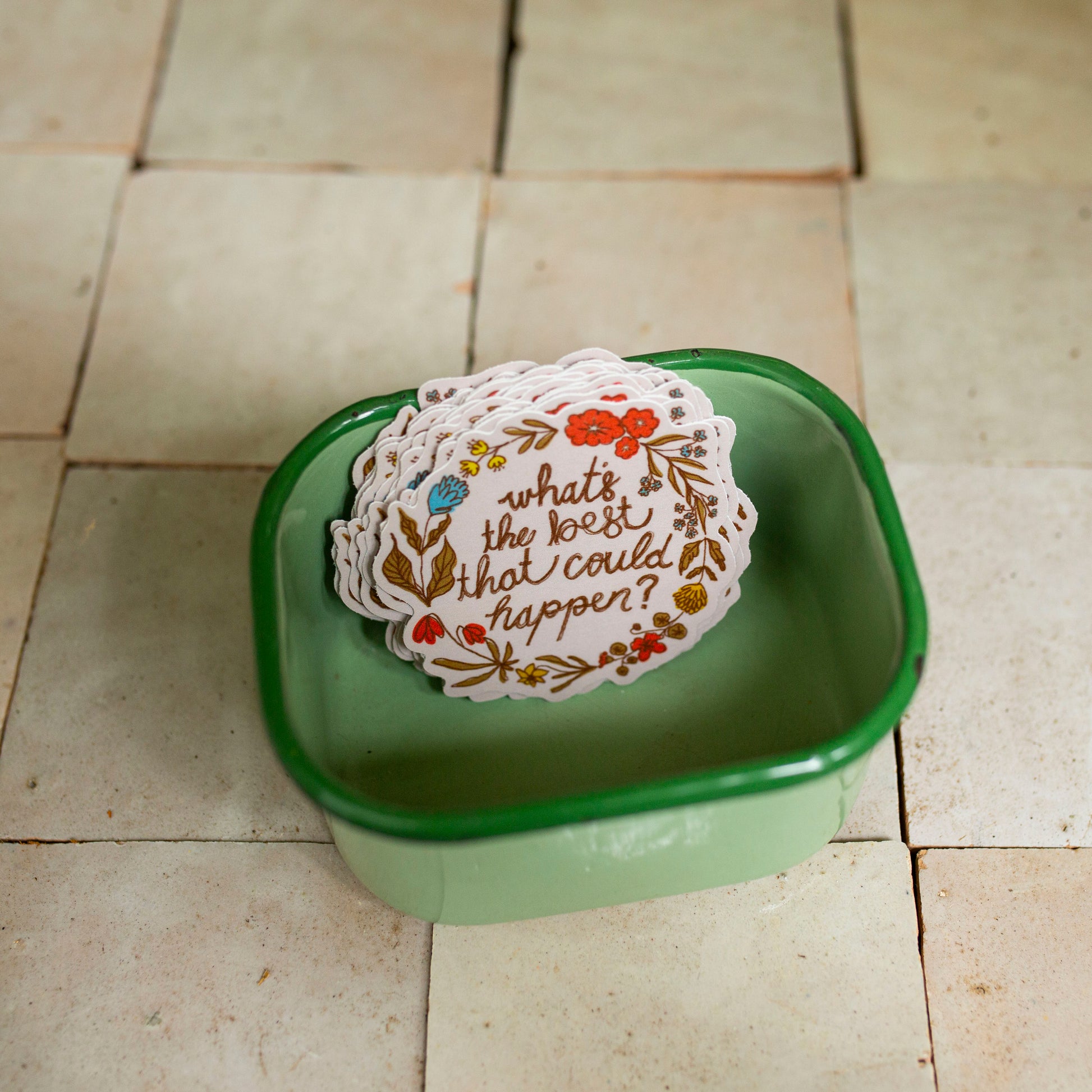 A stack of white paper stickers with floral borders and the phrase 'what's the best that could happen?' printed in brown script, placed inside a small green square dish on a beige tiled floor.