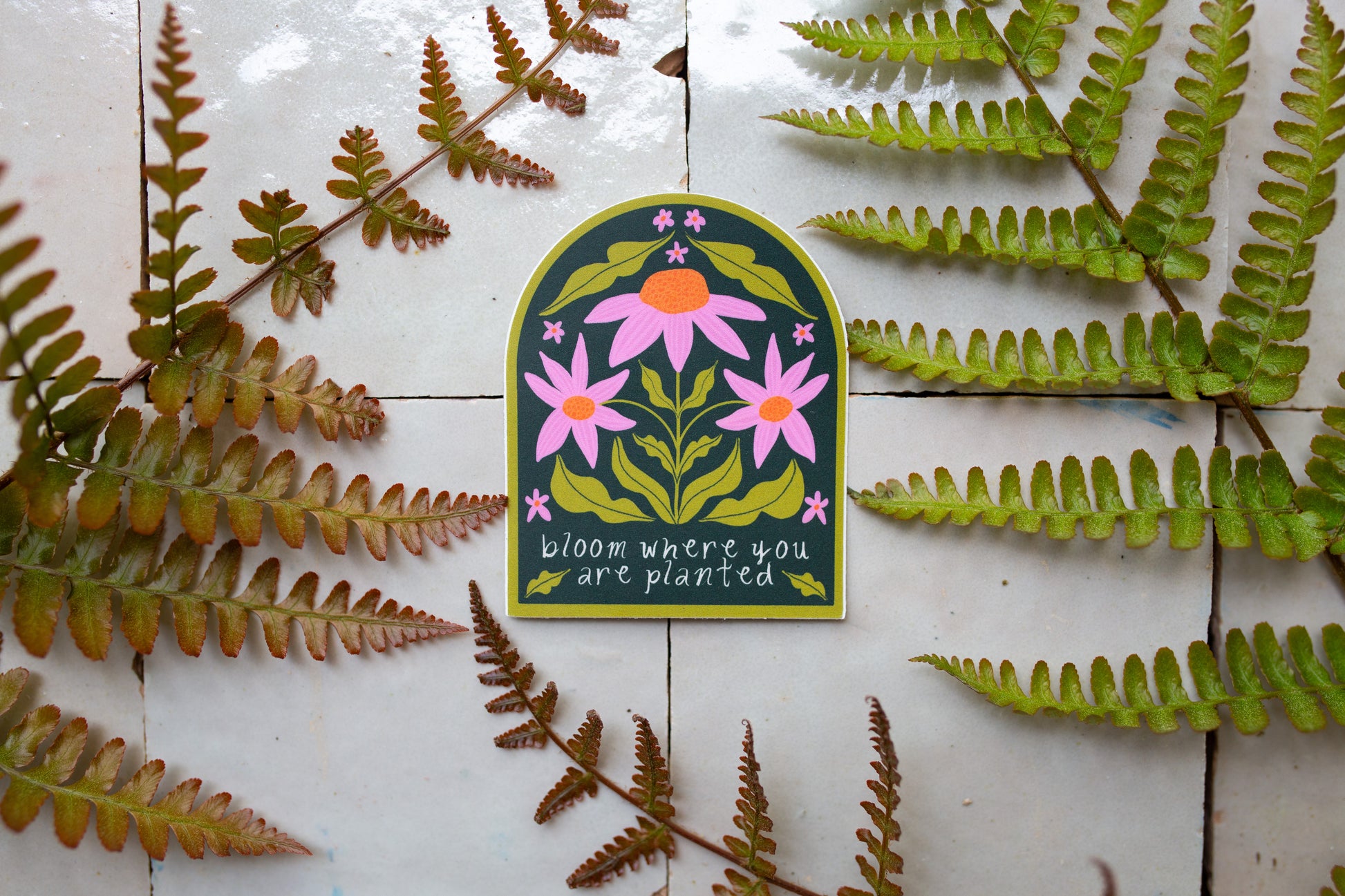 Picture of vinyl sticker that says bloom where you are planted with a dark green background and pink and orange florals.