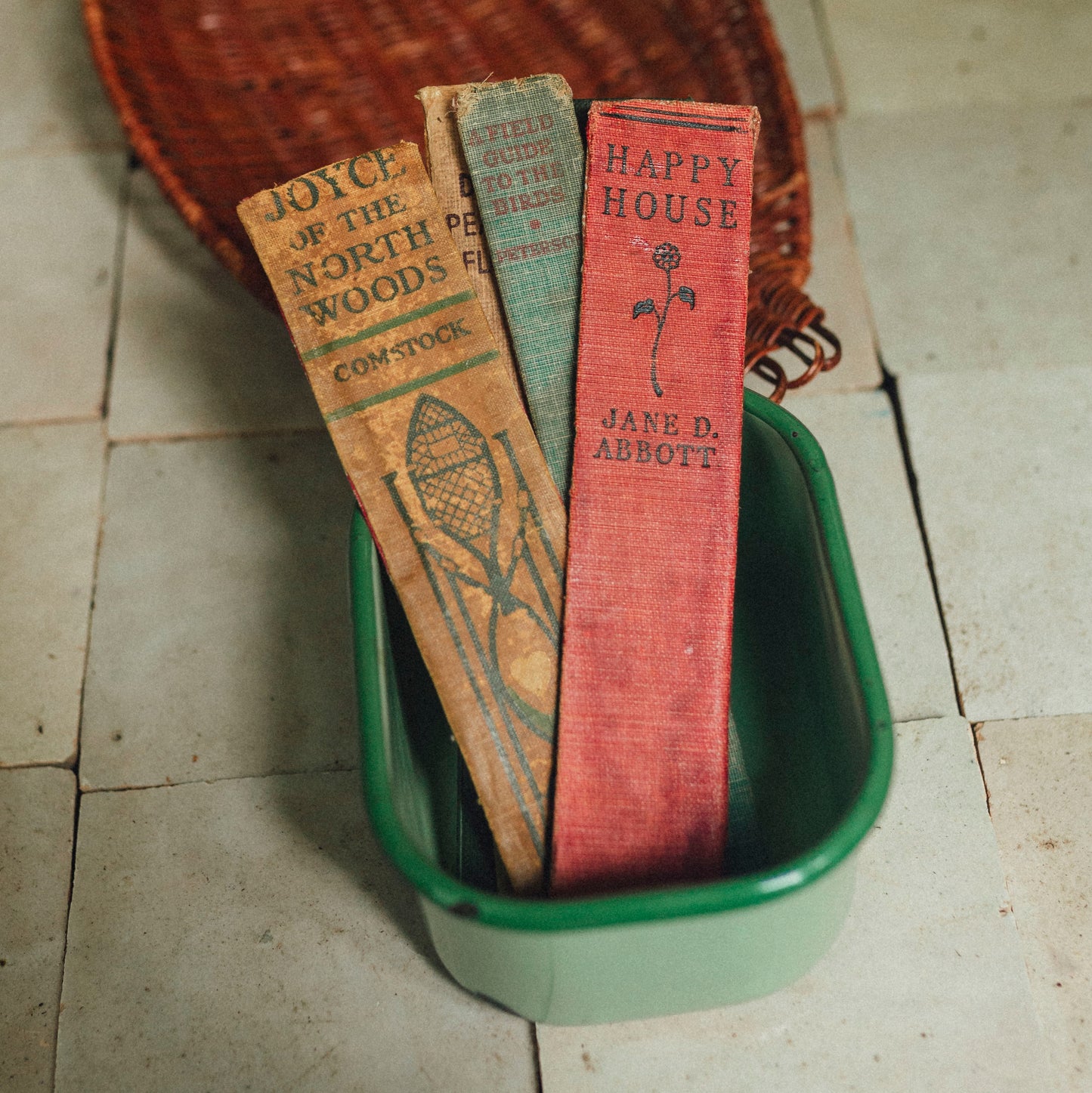 Three vintage book spine bookmarks resting inside a green rectangular dish, with a woven duck-shaped basket behind them on a tiled floor.