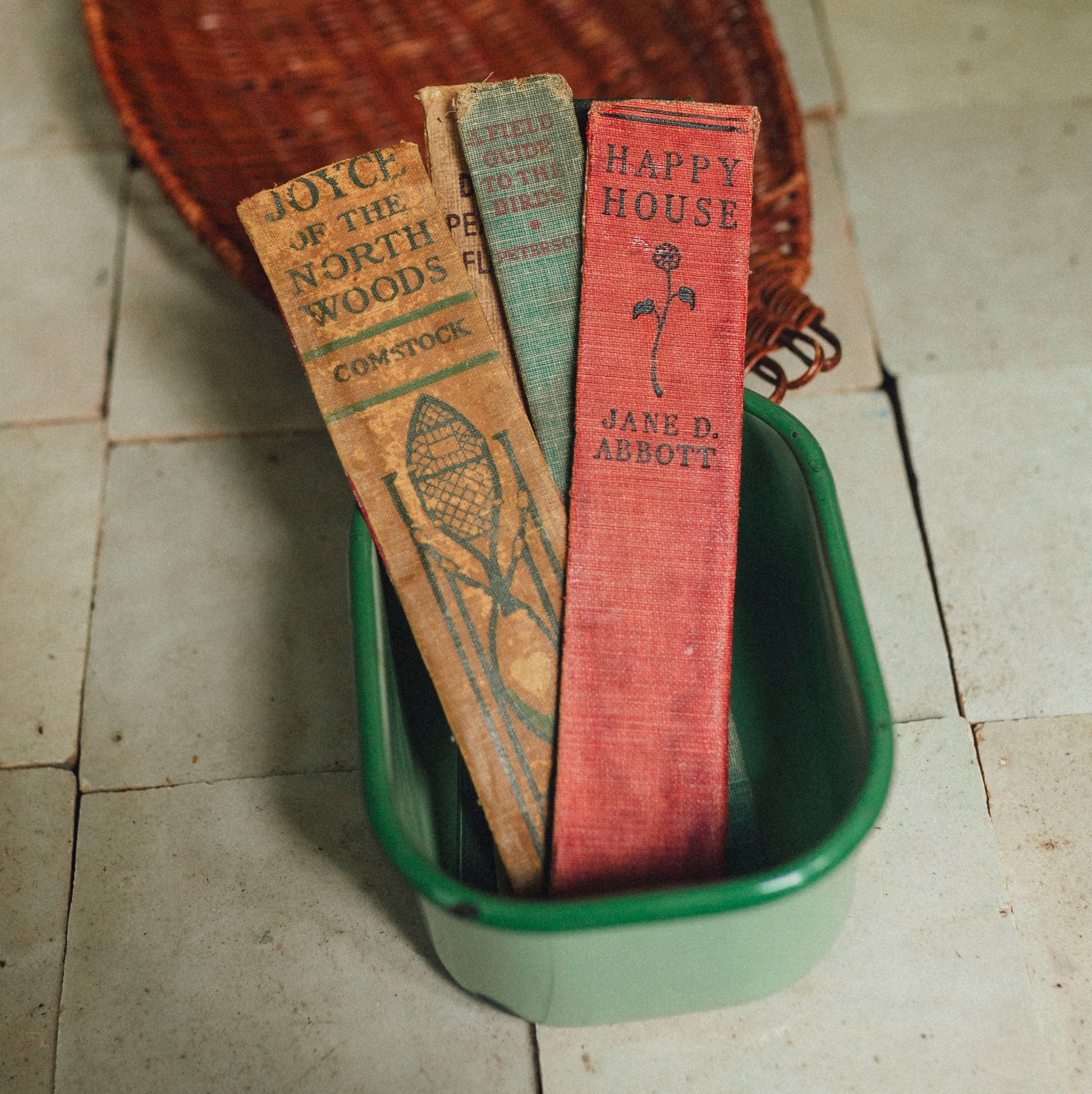 Three vintage book spine bookmarks resting inside a green rectangular dish, with a woven duck-shaped basket behind them on a tiled floor.