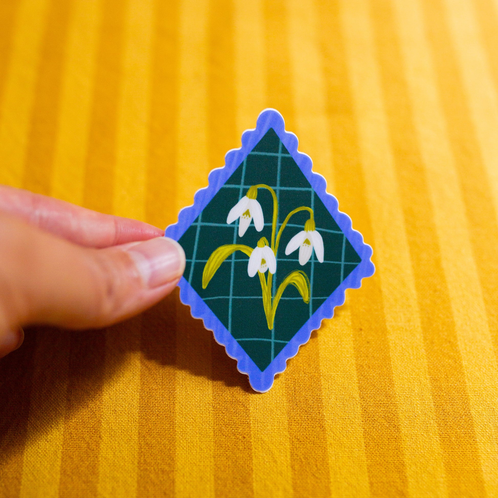 Diamond-shaped sticker with floral design held by a hand on a yellow textured background