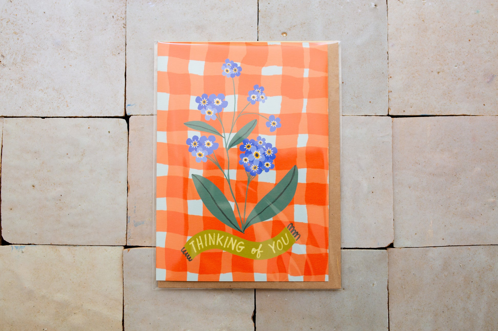 Card floral design and 'Thinking of You' text on an orange checkered background, placed on a stone surface.