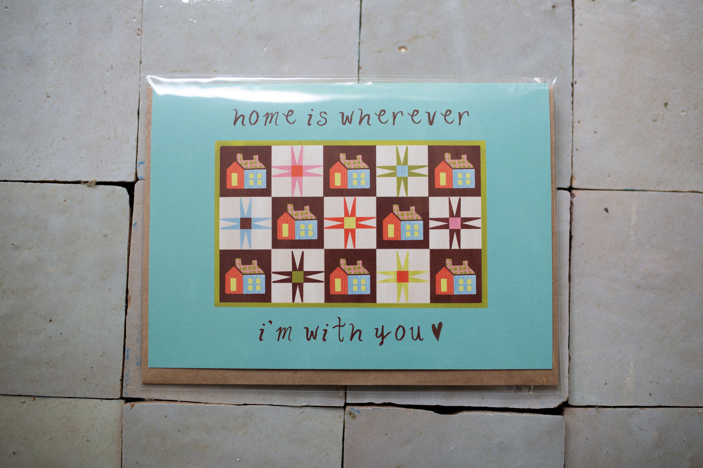 Greeting card with quilt design and text 'home is wherever I'm with you' on a tiled floor.