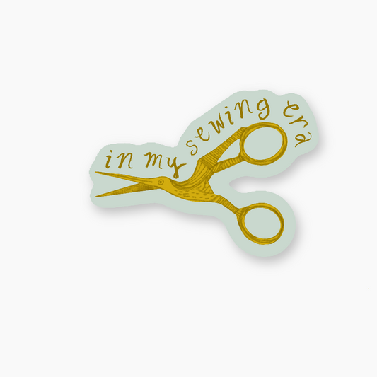 Sticker with gold scissors design and 'in my sewing era' text on a light blue background