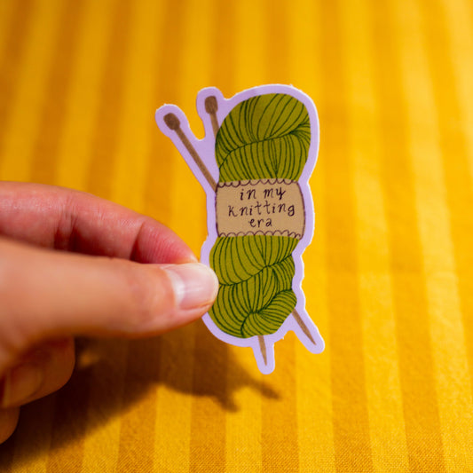 Sticker of a green yarn ball with text "In My Knitting Era" held by a hand on a yellow background