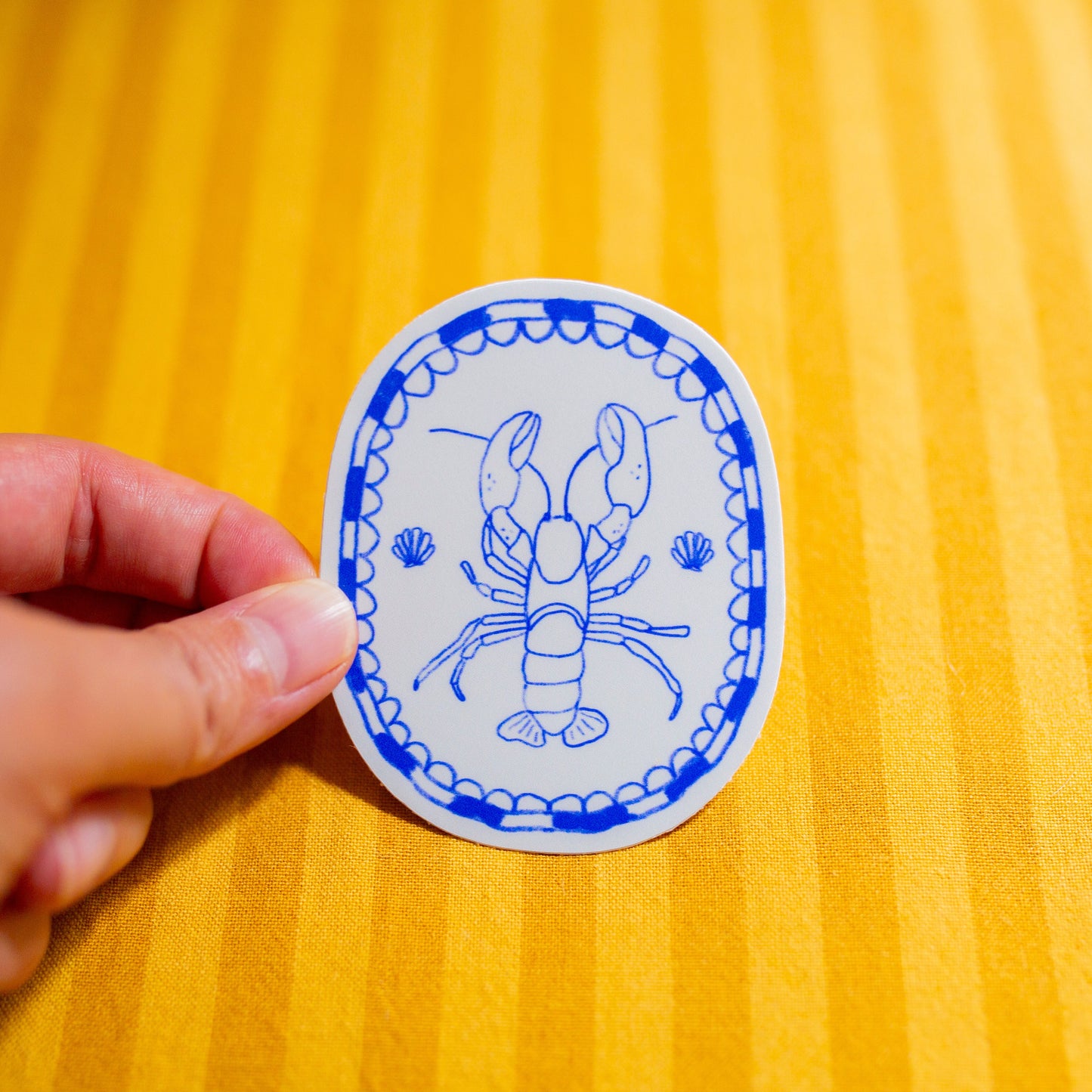 Hand holding a round blue and white sticker with a lobster design on a yellow textured surface.
