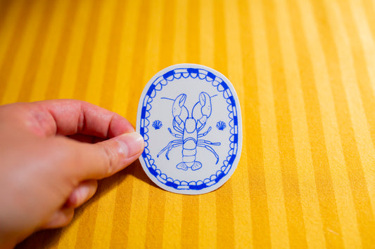 Hand holding a round blue and white sticker with a lobster design on a yellow textured surface.