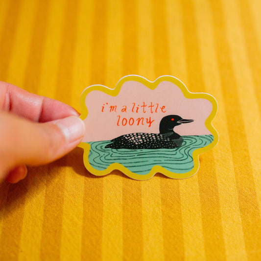 Hand holding a sticker with a loon illustration and text on a yellow background