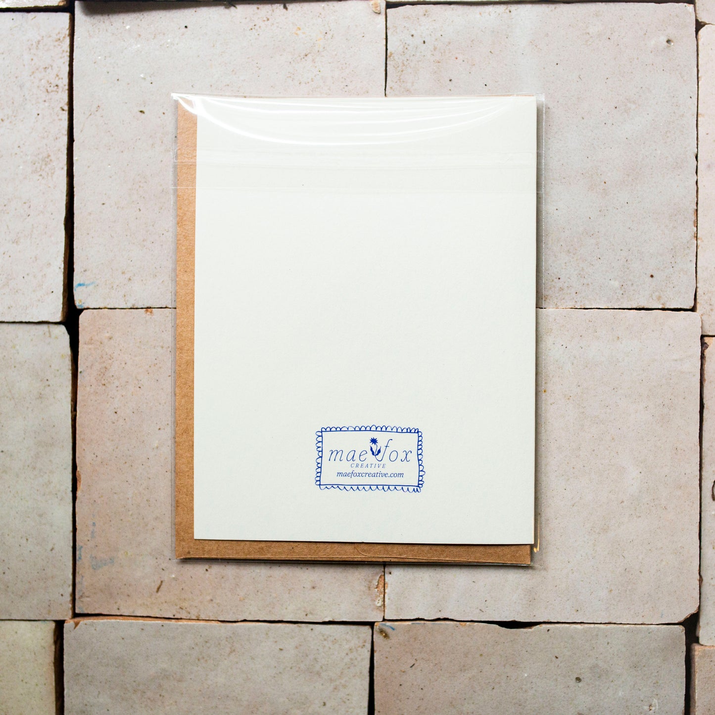 A white stationery card or letterhead with a small blue and white logo at the bottom, placed on top of a brown kraft envelope against a background of light beige stone tiles.
