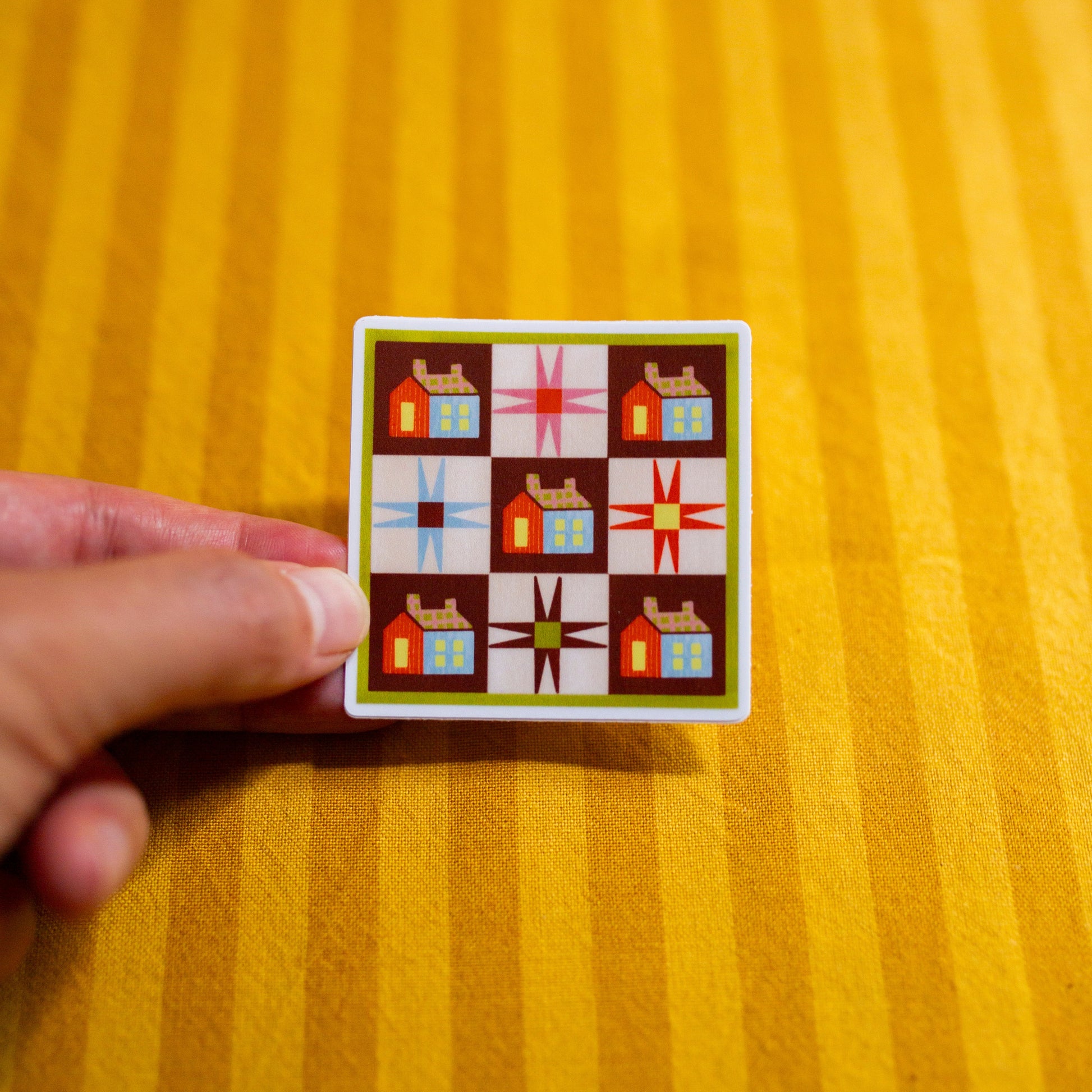 Small sticker with with quilt design held by a hand on a yellow striped background