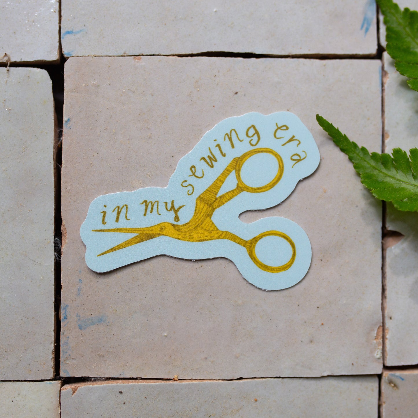 Sticker with scissors and text 'in my sewing era' on a tiled floor with a fern leaf.