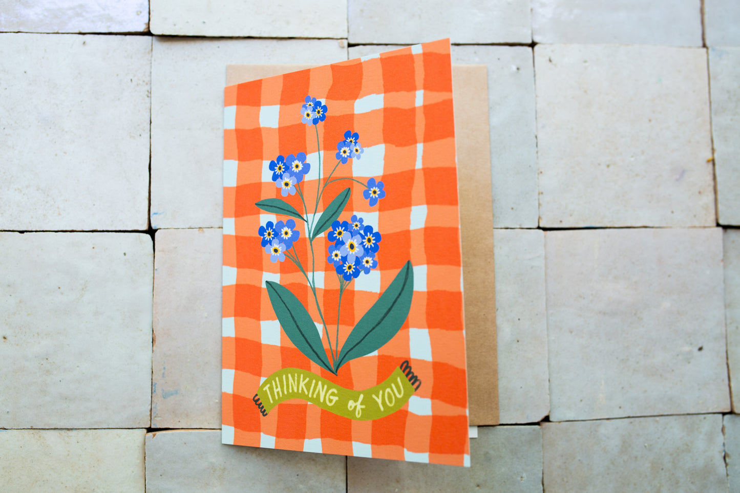 Greeting card with floral design and 'Thinking of You' text on an orange checkered background, placed on a stone surface.