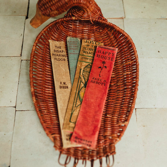 A woven rattan duck-shaped tray holding three vintage book spine bookmarks, placed on a light tiled floor.