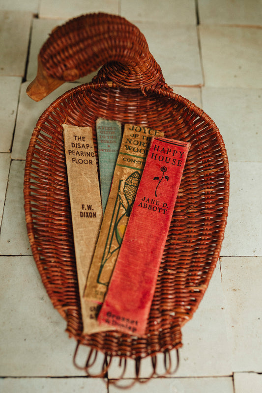 A decorative woven rattan tray shaped like a duck, holding three vintage book spines on a light tile floor.