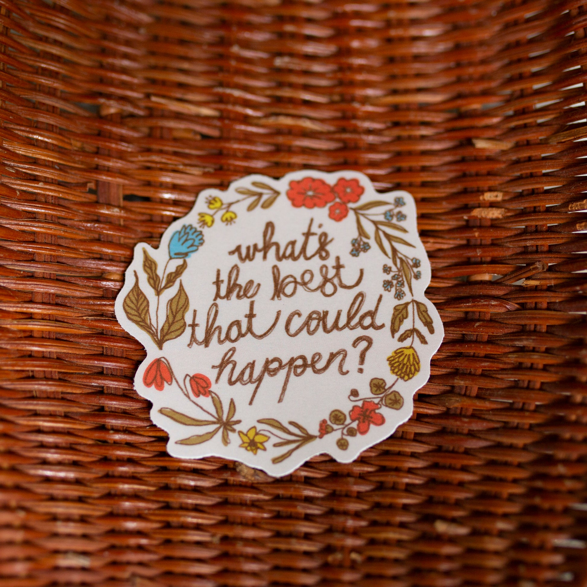 A round white sticker with a floral border and the handwritten text 'what's the best that could happen?' placed inside a brown woven wicker basket on a light tiled floor.