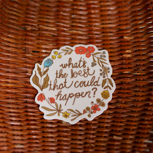 A round white sticker with a floral border and the handwritten text 'what's the best that could happen?' placed inside a brown woven wicker basket on a light tiled floor.