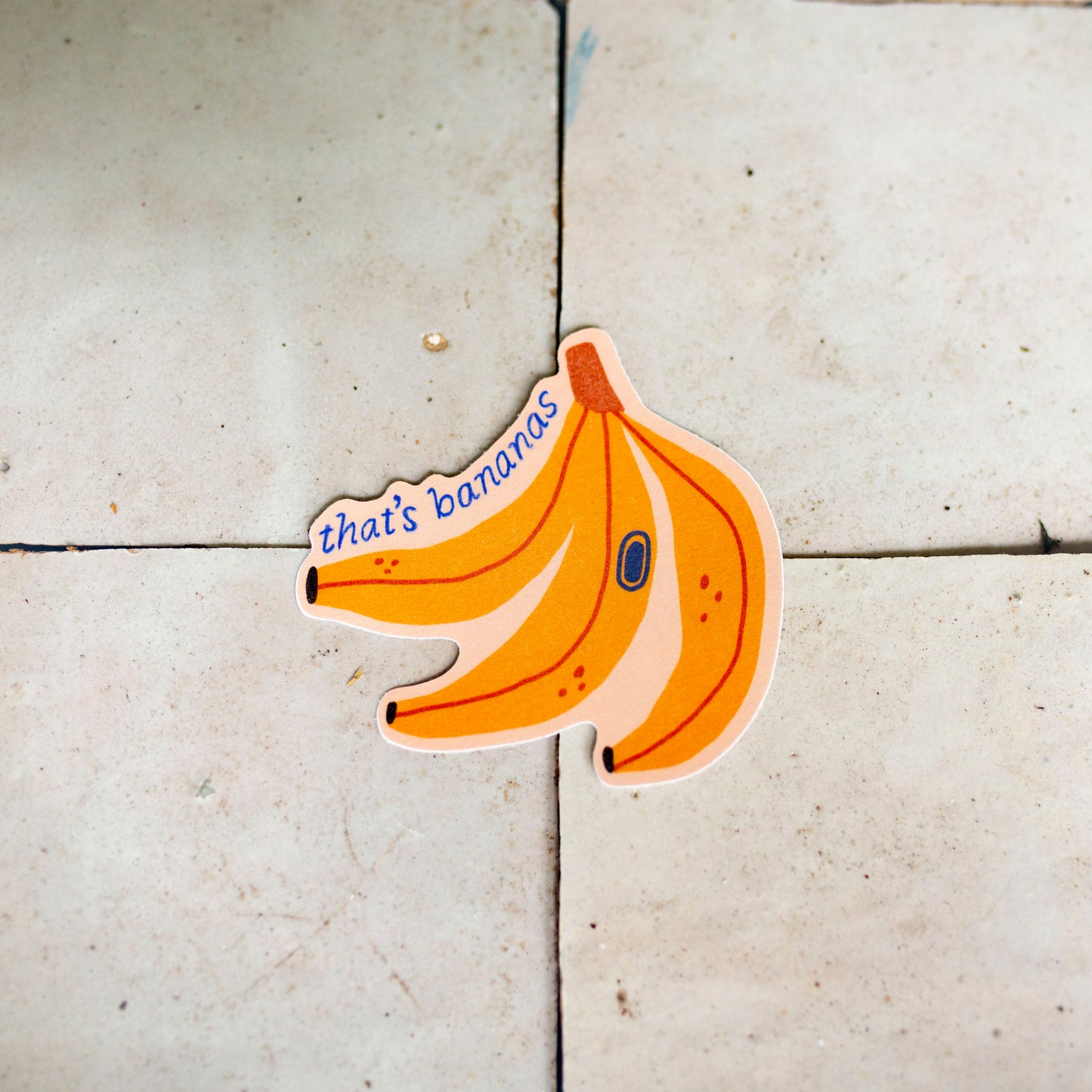 A die-cut sticker shaped like a bunch of three bananas, placed on a light beige tiled floor. The sticker features the text 'that's bananas' in blue script on the left banana, with a small blue oval detail on the middle banana.