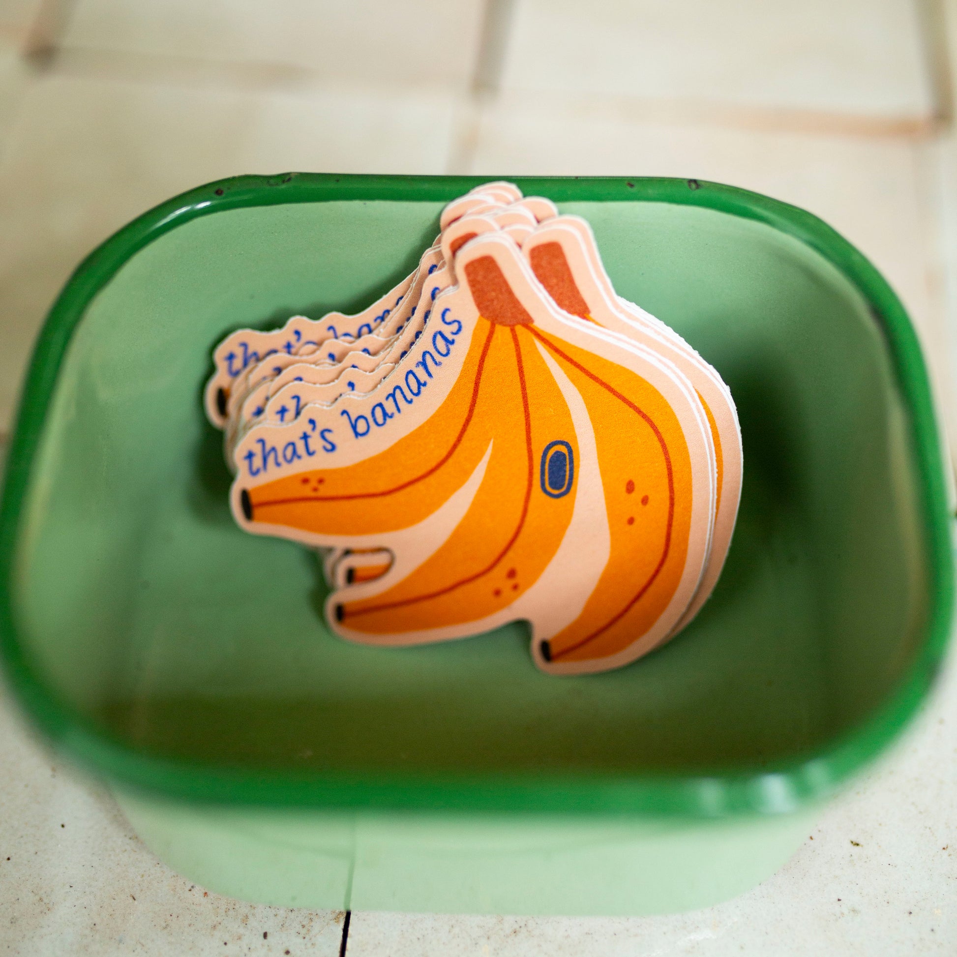 A stack of die-cut banana-shaped magnets or stickers resting inside a green plastic container on a light tiled floor. The top magnet features blue text reading 'that's bananas' 