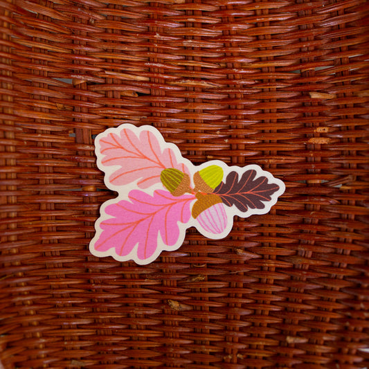 A die-cut sticker featuring pink, brown, and green autumn leaves with acorns, placed on a brown woven wicker basket.