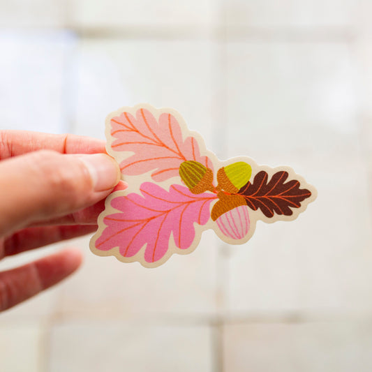A hand holding a small die-cut sticker featuring pink, brown, and yellow autumn leaves and acorns against a softly blurred light background.