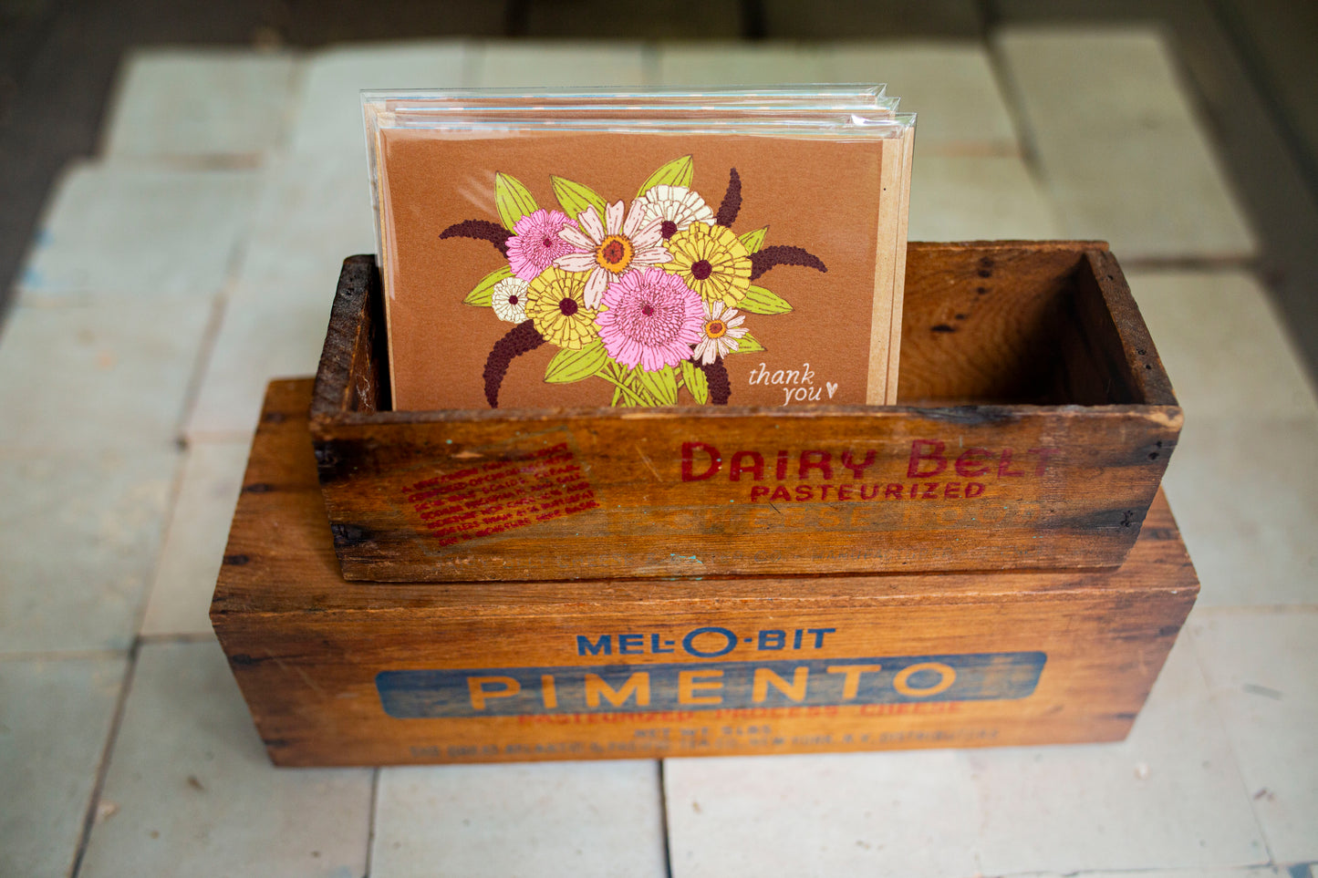 A stack of greeting cards with a floral design and 'thank you' text, displayed in a vintage wooden Melo-bit Pimento box on a tiled floor.
