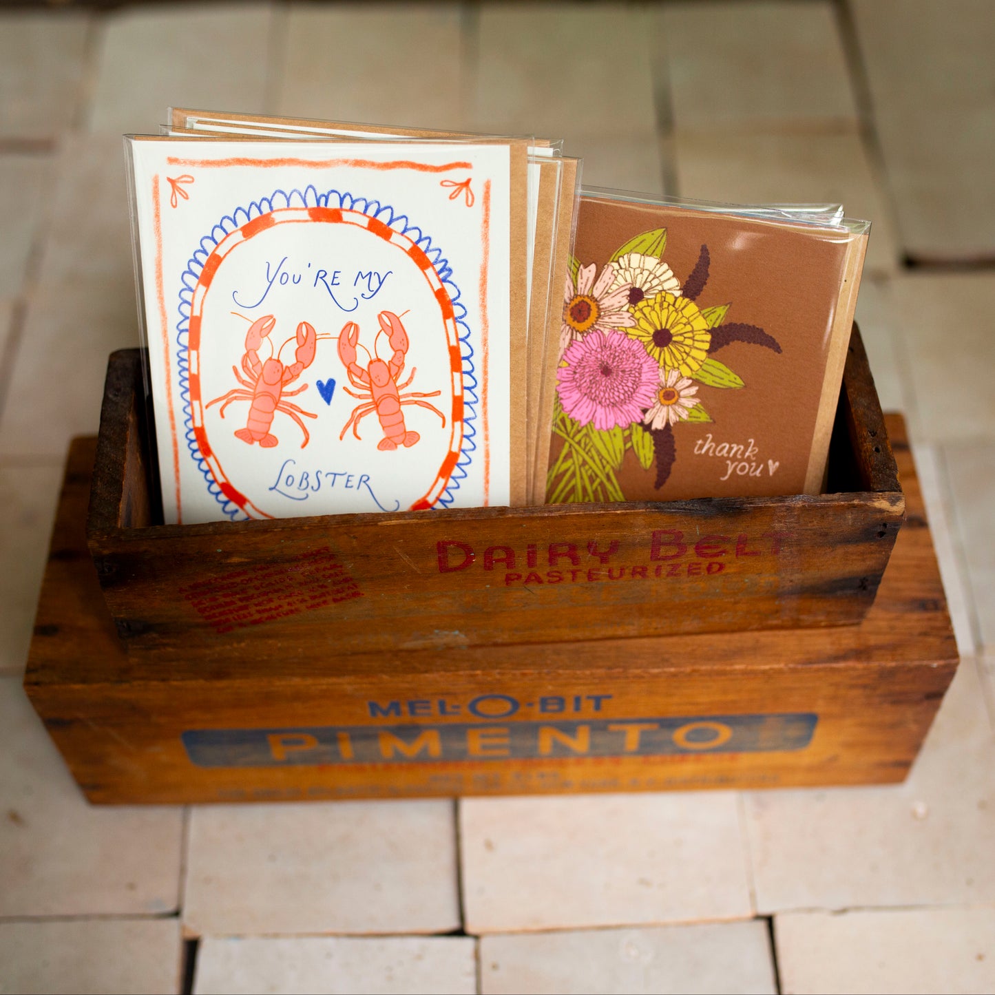 A wooden crate  holding several greeting cards, including one with lobster artwork and another with flowers, placed on a tiled floor.