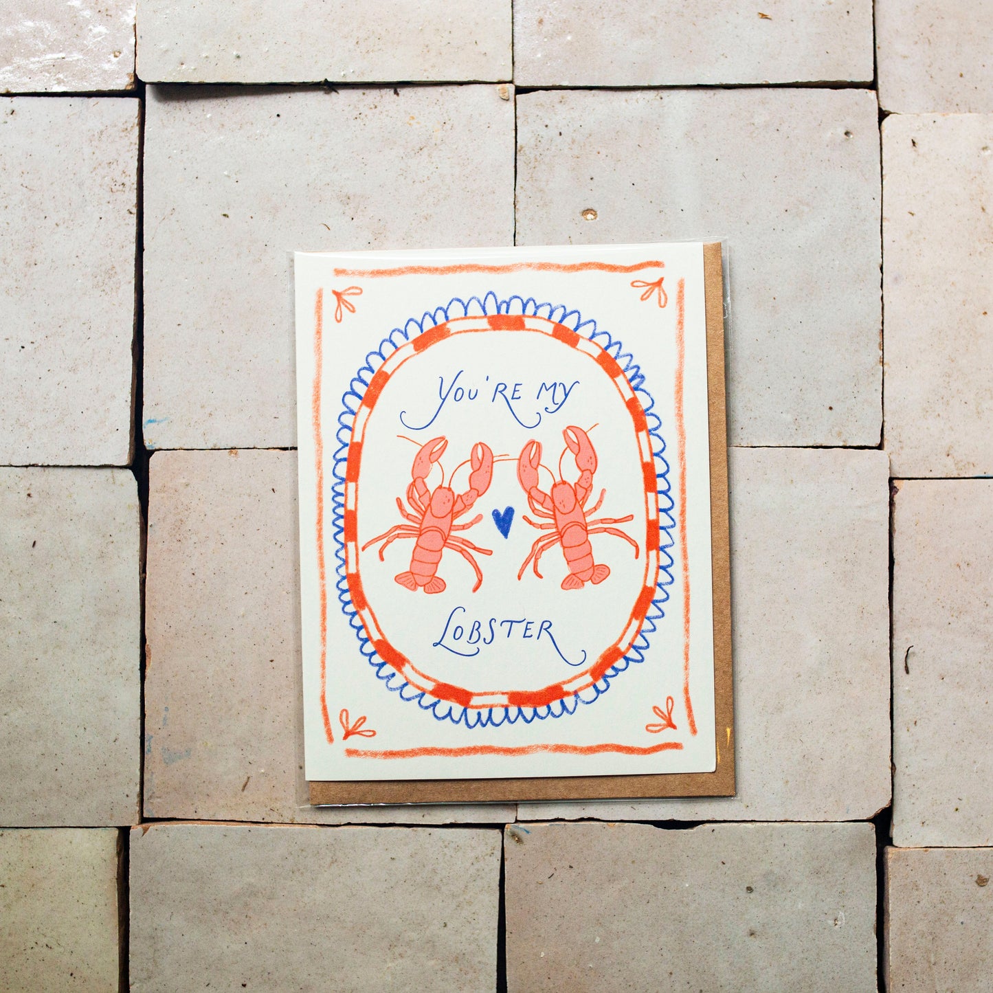A square greeting card featuring a lobster illustration and the text 'You're my Lobster' in blue script, placed against a background of large light beige stone tiles.