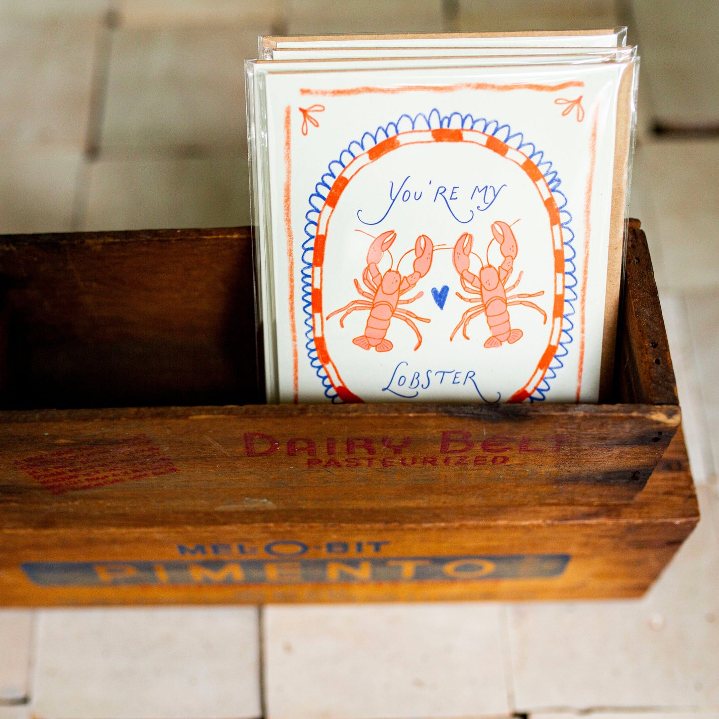 A stack of greeting cards featuring an orange lobster illustration and the text 'You're My Lobster' in blue script, displayed inside a vintage wooden crate on a tiled floor.