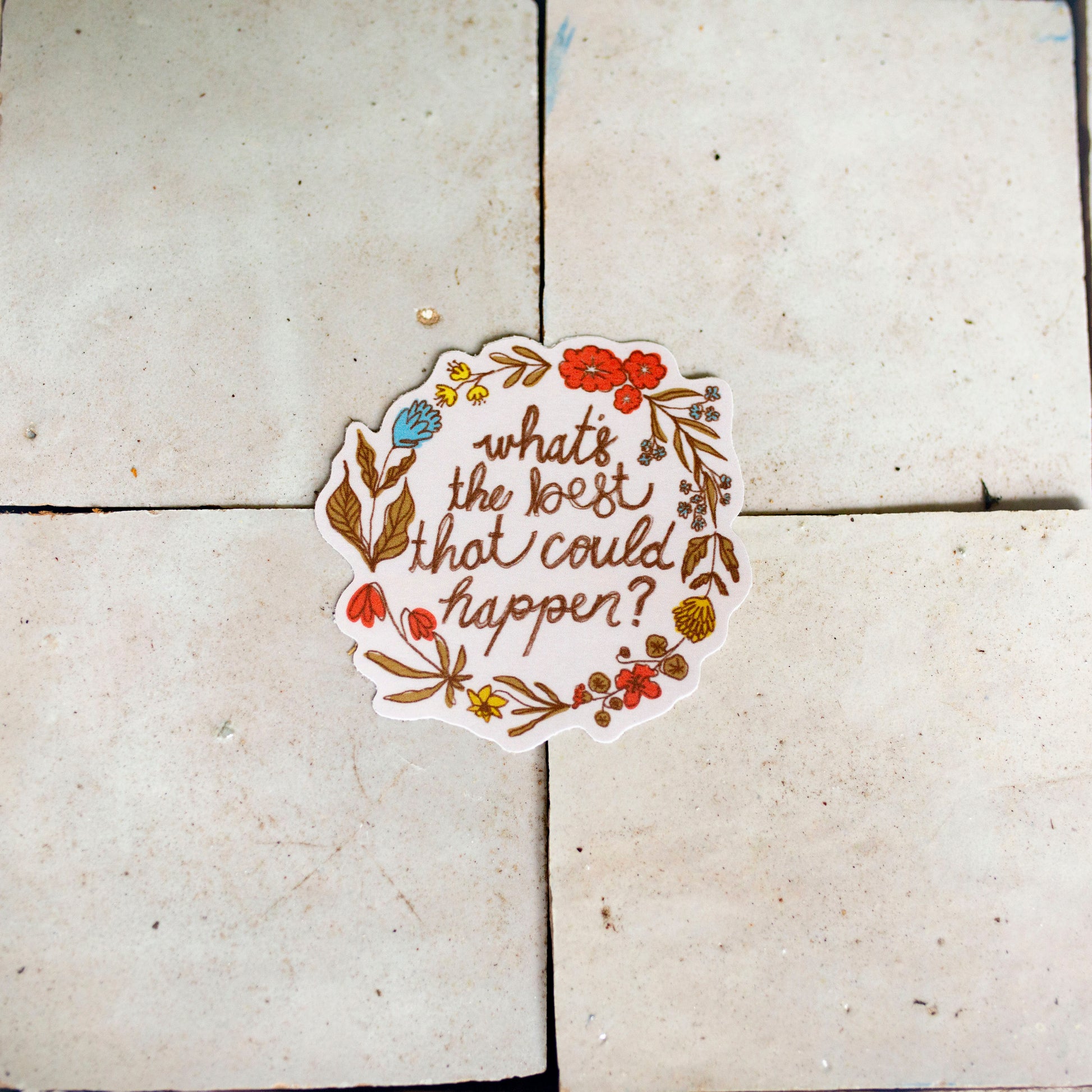 A die-cut sticker with the phrase 'what's the most that could happen?' in brown script, surrounded by a colorful floral wreath, placed on a light beige tiled surface.
