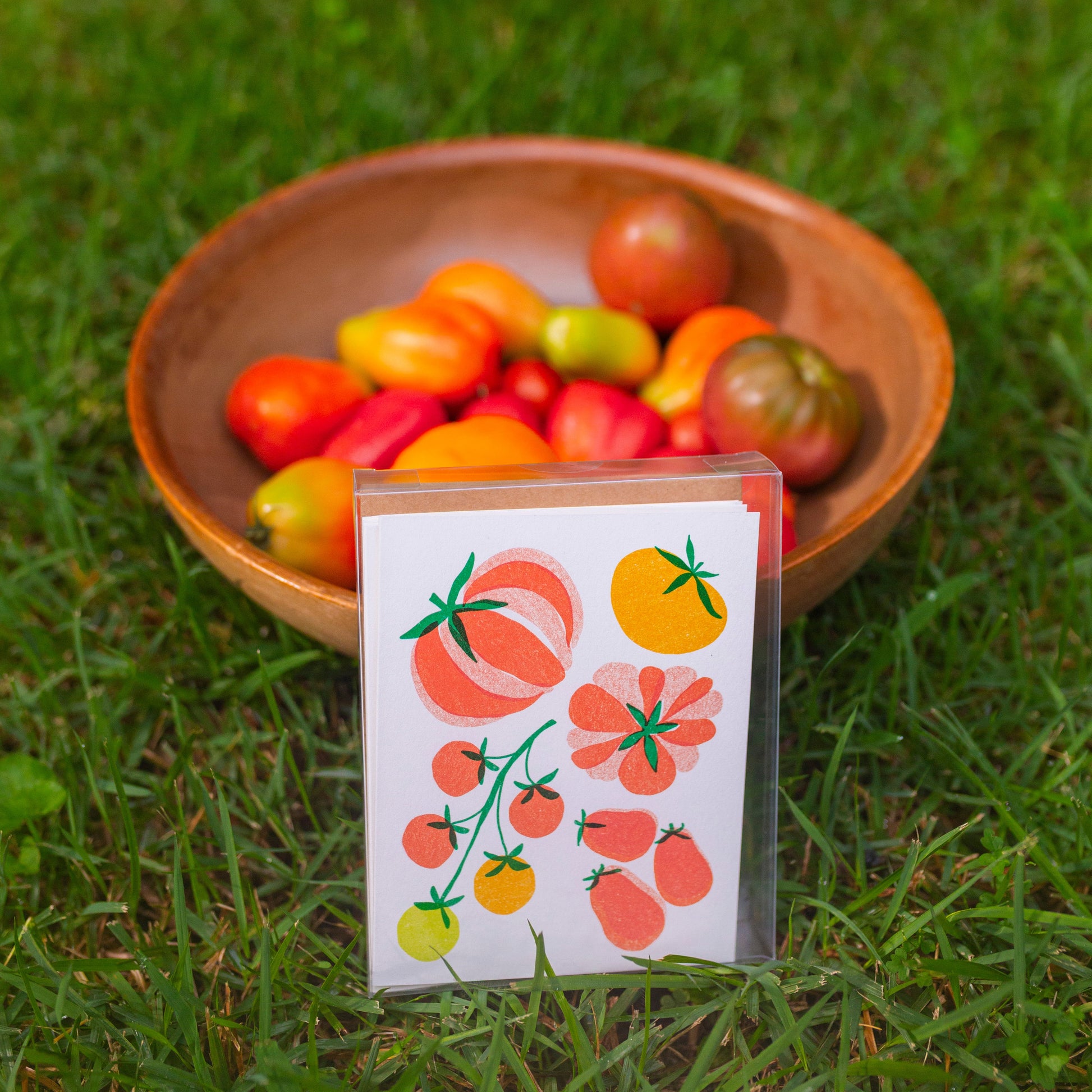 A small printed card with tomato illustrations stands in front of a wooden bowl filled with assorted tomatoes on a green grass background.
