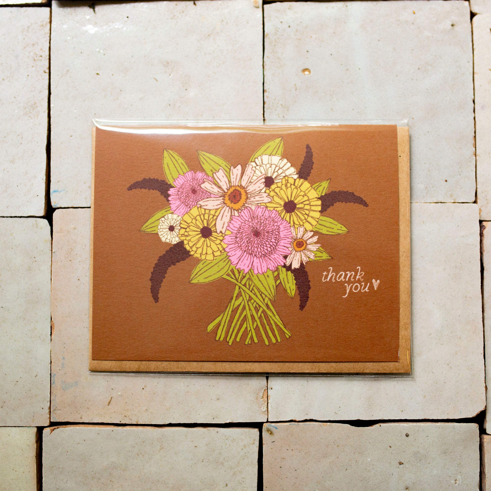 A square brown greeting card featuring a colorful bouquet of pink, yellow, and white flowers with green leaves, placed on a light beige tiled surface. The card is printed with 'thank you' in white script on the bottom right.