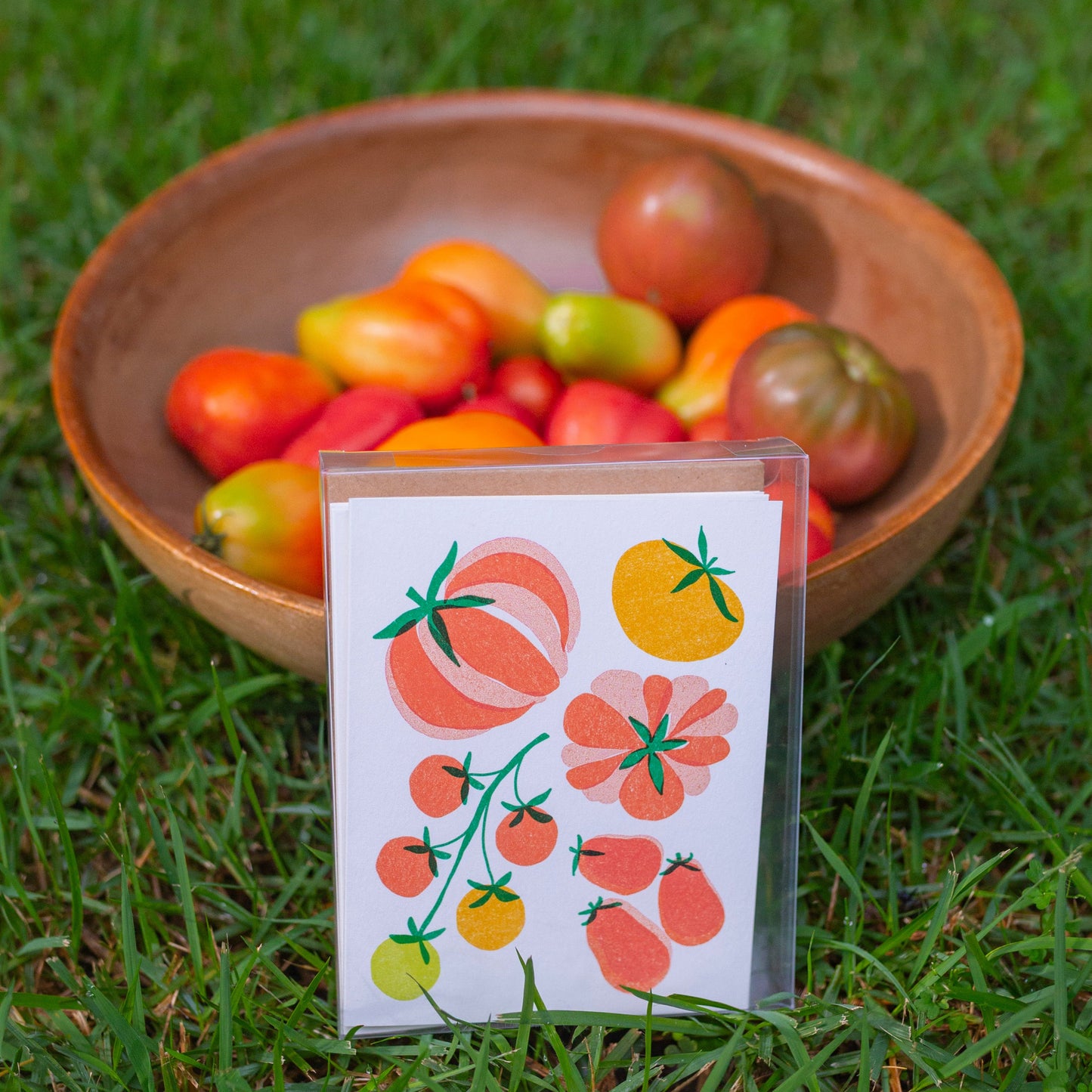 A small printed card with tomato illustrations stands in front of a wooden bowl filled with assorted tomatoes on a grassy lawn.