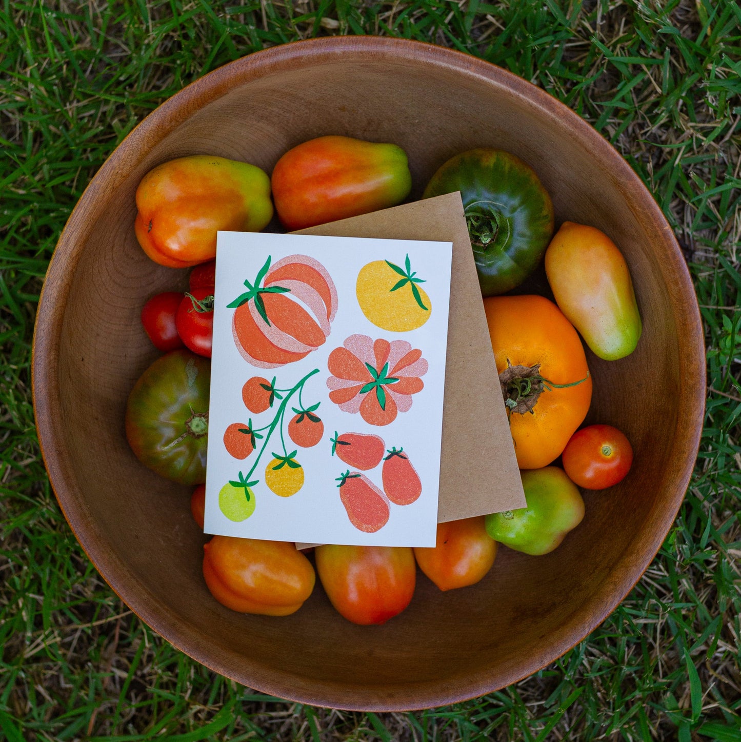 A greeting card featuring colorful tomato illustrations, placed on top of a brown kraft envelope inside a rustic  bowl filled with fresh tomatoes, set on a green grass lawn.